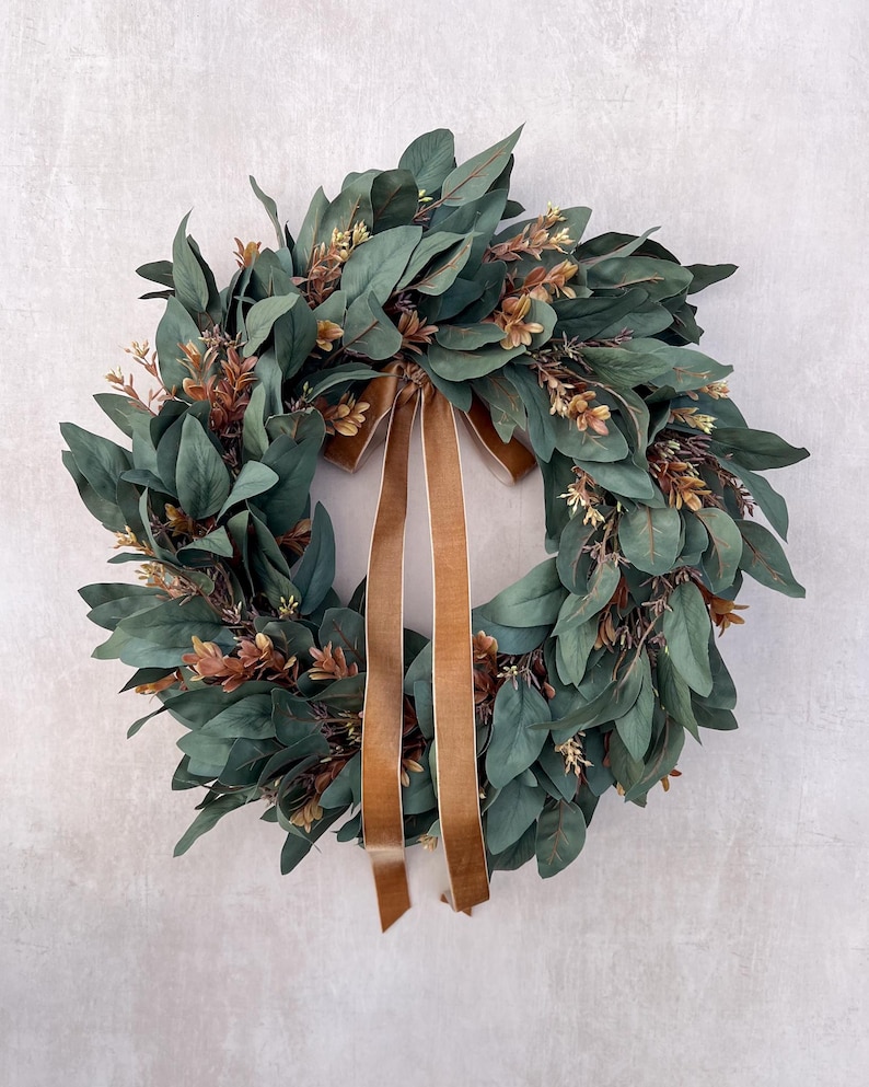 May include: A circular wreath constructed from dark green eucalyptus leaves and interspersed with small brown sprigs. A long, smooth brown velvet ribbon is tied at the top center, with two tails hanging down through the middle of the wreath.