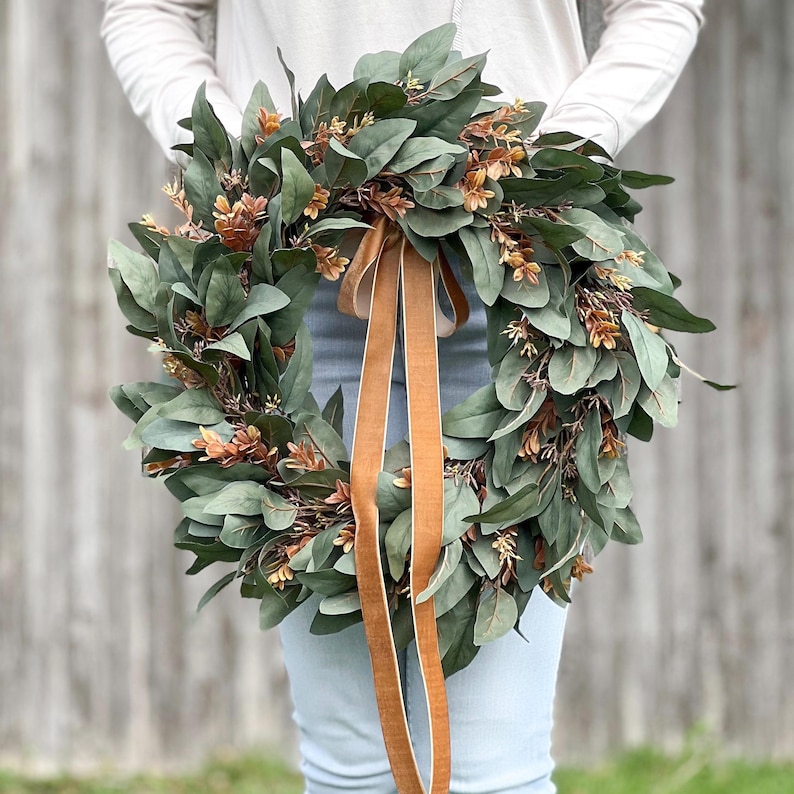 May include: A circular wreath composed of dark green eucalyptus leaves and interspersed with small, copper-toned foliage accents. A long, smooth, tan-colored ribbon hangs vertically from the top center of the wreath, draping downward.
