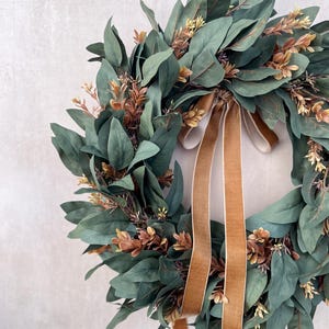 May include: A circular wreath composed of dark green eucalyptus leaves and small clusters of brownish-orange foliage. A long, smooth, tan-colored velvet ribbon is tied in a bow at the top, with two long tails hanging down through the center.