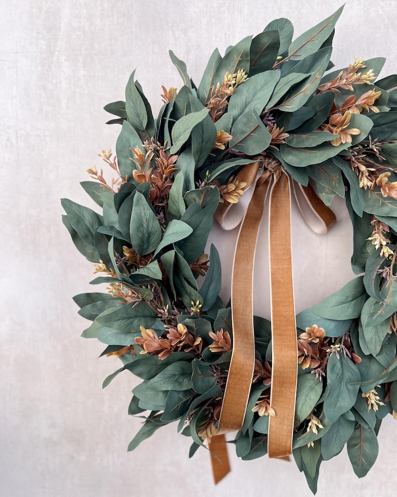 May include: A circular wreath composed of dark green eucalyptus leaves and small clusters of brownish-orange foliage. A long, smooth, tan-colored velvet ribbon is tied in a bow at the top, with two long tails hanging down through the center.