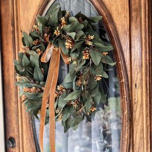 May include: A circular wreath featuring dark green leaves and small light brown floral accents. The wreath is adorned with two long, smooth, tan-colored velvet ribbons that hang vertically from the top center, creating a decorative display.