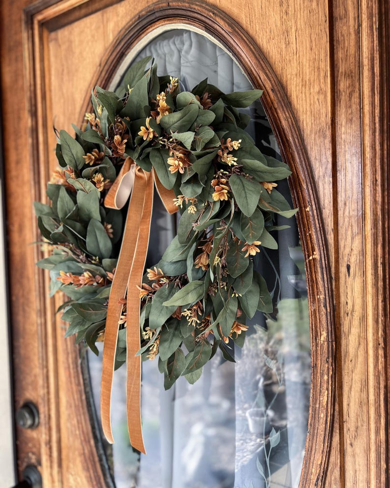 May include: A circular wreath featuring dark green leaves and small light brown floral accents. The wreath is adorned with two long, smooth, tan-colored velvet ribbons that hang vertically from the top center, creating a decorative display.