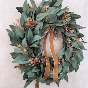 May include: A circular wreath constructed from dense, dark green eucalyptus leaves interspersed with small clusters of light brown and tan foliage. Two long, smooth, golden-brown velvet ribbons hang vertically from the center of the wreath.