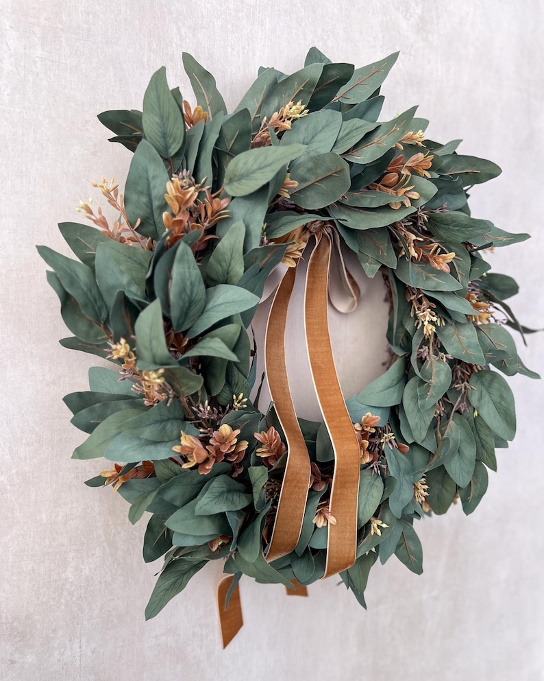 May include: A circular wreath constructed from dense, dark green eucalyptus leaves interspersed with small clusters of light brown and tan foliage. Two long, smooth, golden-brown velvet ribbons hang vertically from the center of the wreath.