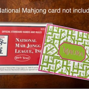 May include: Two cards on a wooden surface. One is red with white text, including "National Mah Jongg League, Inc." and "2026". The other is white with a green bamboo pattern and the name "MAURA" in a green octagon.