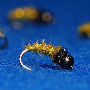May include: Close-up of a fishing fly with a curved hook. The fly has a dark body and a green, textured head. The hook is silver, and the fly is set against a blue background.