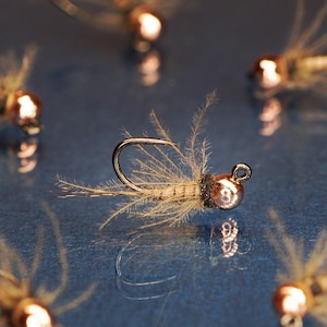 May include: Close-up of a copper-colored fly fishing lure with a brown and white body and a feathery tail. The lure is on a blue surface.