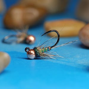 May include: Close-up of a fishing fly with a black hook, a silver and copper-colored bead, and green and brown accents. The fly is set against a bright blue background, with blurred pebbles in the background.