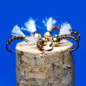 May include: Close-up of several fishing flies with gold-colored heads, brown bodies, and white feather wings. The flies are arranged on a cork surface against a bright blue background. The hooks are black.
