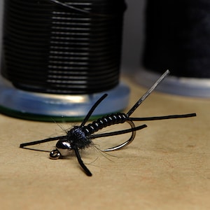 May include: A black fly fishing lure with a segmented body and black feathers. The lure is tied on a hook and is ready to be used for fishing.