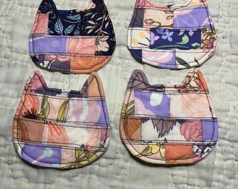 May include: Four handmade cat-shaped coasters. Each coaster is made of patchwork fabric with floral and geometric patterns in shades of pink, purple, orange, and navy blue. The coasters have a quilted appearance and are arranged on a white, textured surface.