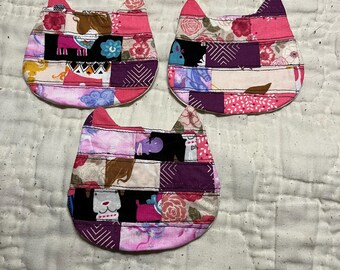 May include: Three handmade cat-shaped coasters. Each coaster is made from fabric scraps in shades of pink, purple, and black, with floral and geometric patterns. The coasters are arranged on a white quilted surface.