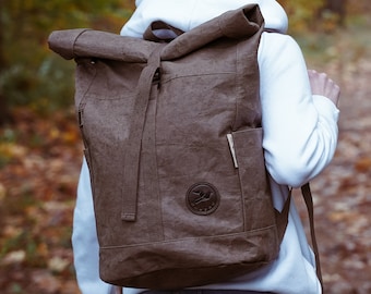 Könnte beinhalten: Eine Person trägt eine weiße Strickmütze und ein weißes Hoodie mit einem braunen Canvas-Rucksack. Der Rucksack hat einen Rollverschluss und ein Logo auf der Vorderseite.
