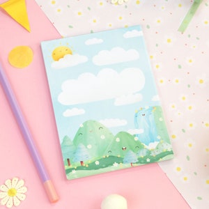 May include: A notepad with a whimsical landscape illustration featuring a smiling sun, clouds, and green hills. A purple pencil, decorative flowers, and colourful accents surround the notepad on a pink surface.