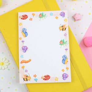 Może przedstawiać: Biały notes z różowym obramowaniem i kolorowym obramowaniem z owadami. Obrzeże przedstawia motyle, biedronki, pszczoły i gąsienice. Notes znajduje się na żółtym i różowym tle.