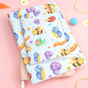 May include: Two blue notebooks with a colorful pattern of bees, butterflies, ladybugs, caterpillars, and flowers. The notebooks are stacked on top of each other and are sitting on a pink surface.