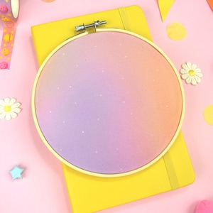 Pode incluir: Um bastidor redondo com um design de gradiente pastel em tons de rosa, laranja e roxo, salpicado de pontos brancos. O bastidor repousa sobre um caderno amarelo, rodeado por elementos decorativos como fita washi, bolas de feltro e enfeites em forma de estrela.