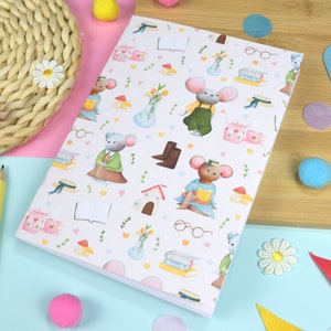 Pode incluir: Um caderno branco com um padrão repetitivo de ratos, livros, óculos, flores e casas. O caderno está sobre uma superfície azul claro com uma superfície amarela e rosa atrás.