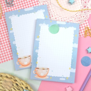 Pode incluir: Dois blocos de notas azuis claros com um padrão de grade e uma borda de nuvens e estrelas. Cada bloco de notas apresenta uma ilustração de xícara de chá de desenho animado. Os blocos são cercados por itens decorativos em uma superfície rosa.