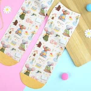Könnte beinhalten: Ein Paar cremefarbene Socken mit einem sich wiederholenden Muster von Mäusen, die Brillen tragen und Bücher halten. Die Socken haben gelbe Bündchen und liegen auf einem rosa und blauen Hintergrund.