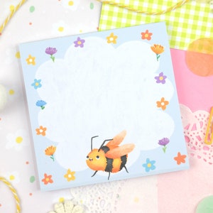 May include: A blue notepad with a white cloud and a cartoon bee with black stripes and orange and yellow fur. The cloud is surrounded by colorful flowers.