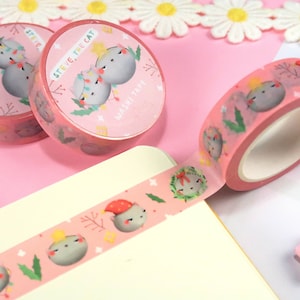 May include: Pink washi tape rolls featuring a cartoon cat character with festive lights and the text "STEVE, THE CAT". The tape is decorated with repeating cat illustrations, holly, and stars. The tape is unrolled on a white surface.