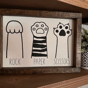 May include: A white wooden sign with black paw prints and the words "Rock, Paper, Scissors".
