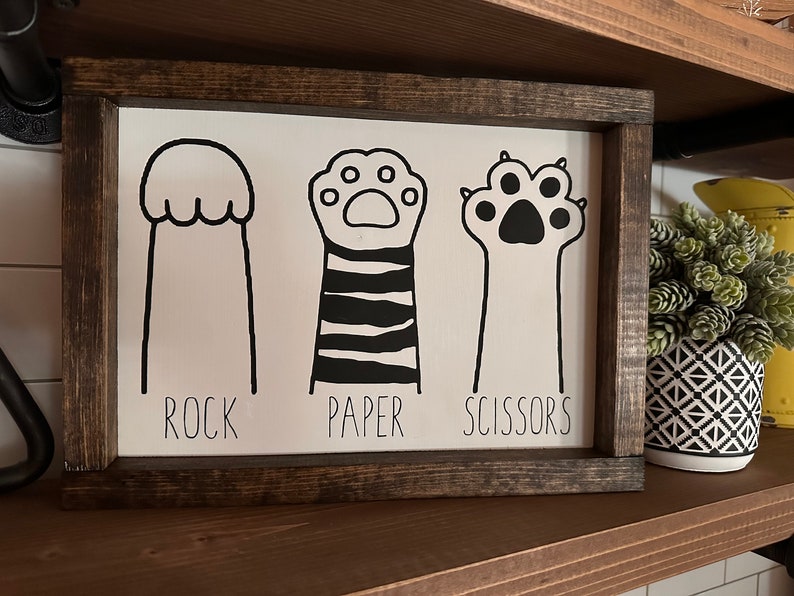 May include: A white wooden sign with black paw prints and the words "Rock, Paper, Scissors".