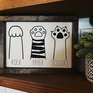 May include: Black and white sign with a paw print, a paw print with stripes, and a paw print with claws. The sign says "Rock, Paper, Scissors".