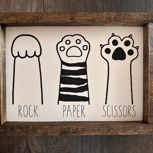 May include: A white wooden sign with black illustrations of a cat paw, a cat paw with stripes, and a cat paw with claws. The words "ROCK", "PAPER", and "SCISSORS" are written below the illustrations.