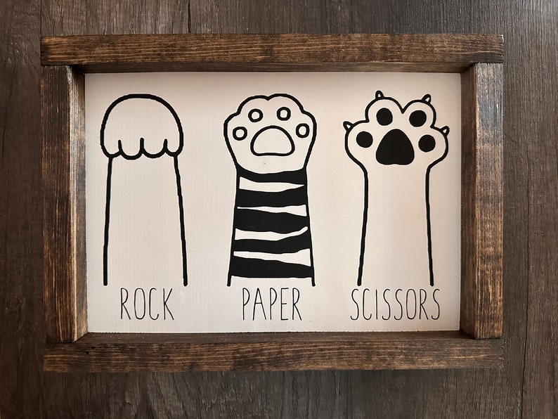 May include: A white wooden sign with black illustrations of a cat paw, a cat paw with stripes, and a cat paw with claws. The words "ROCK", "PAPER", and "SCISSORS" are written below the illustrations.