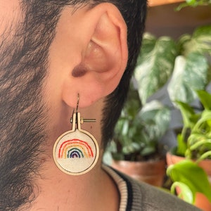 Peut inclure: Boucles d'oreilles arc-en-ciel brodées. Chaque boucle d'oreille présente un petit cercle de broderie en bois avec un motif arc-en-ciel coloré brodé sur du tissu blanc. Les boucles d'oreilles ont un crochet doré.