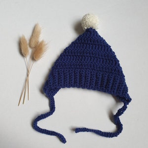 May include: A blue crocheted hat with a white pom pom on top. The hat has two long ties that can be tied under the chin.