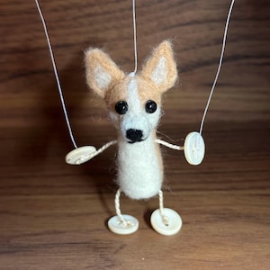 May include: A handmade felt dog puppet with tan and white coloring. The dog has large black eyes, button feet, and button hands. The puppet is suspended by white strings, giving the appearance of a marionette. The background is a warm brown.