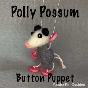 May include: A needle-felted possum puppet with a white face, black ears, and a grey body. It features pink button hands and feet, a pink nose, and black bead eyes. Thin white strings are attached to the limbs. Text reads: Polly Possum Button Puppet Poodie Pin Cushion.