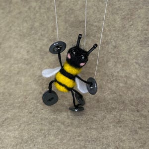 May include: A handmade felt bee puppet with black and yellow stripes, white wings, and button accents. The bee is suspended by strings, giving the appearance of flight. The puppet has a cheerful expression with pink cheeks and is a whimsical craft item.