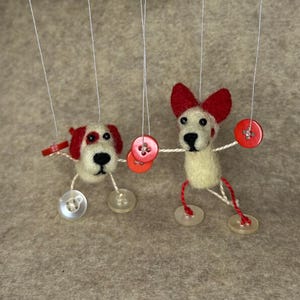 May include: Handmade felt dog puppets with red and white accents. The puppets are suspended by strings and feature red button details. The dogs have button feet and are set against a neutral background.
