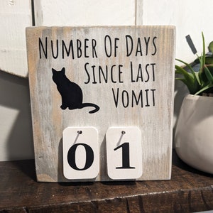 May include: White wooden sign with black text that reads "Number of Days Since Last Vomit". A black silhouette of a cat is above the text. The sign has two white flip cards with black numbers "01" on them.