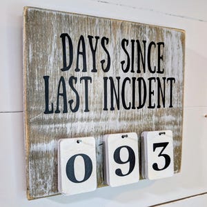 May include: A rustic wooden sign with the text "DAYS SINCE LAST INCIDENT" in black lettering. Below the text are three small, white number cards displaying the numbers "093". The sign is mounted on a white wooden wall.