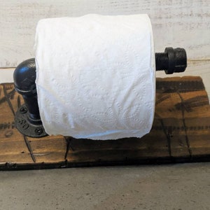 May include: A rustic toilet paper holder made from reclaimed wood and black pipe fittings. The holder is mounted on a wooden base and has a roll of toilet paper on it.
