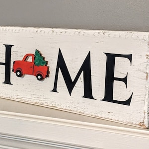 May include: A rectangular wooden sign with the word "HOME" in large black letters. A red truck carrying a green Christmas tree is on the left side. The sign has a distressed, white-painted finish, suggesting a rustic or farmhouse style.