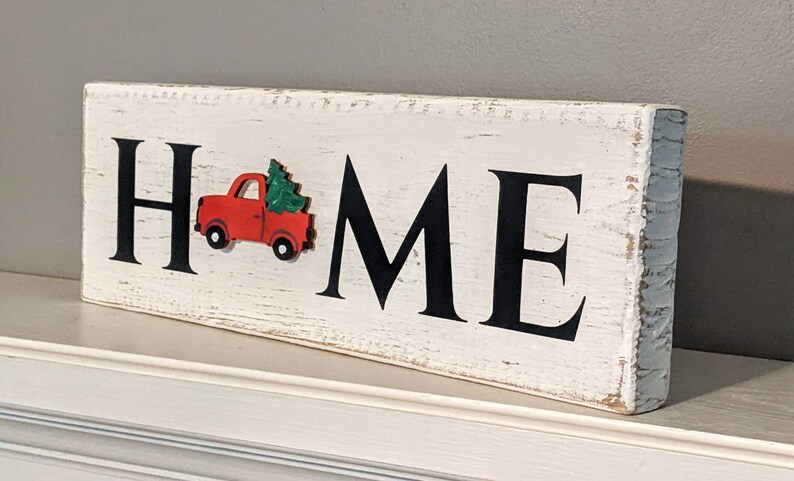 May include: A rectangular wooden sign with the word "HOME" in large black letters. A red truck carrying a green Christmas tree is on the left side. The sign has a distressed, white-painted finish, suggesting a rustic or farmhouse style.