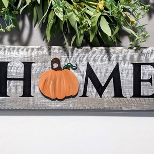 May include: A rustic wooden sign with the word "HOME" in black letters. An orange pumpkin is affixed to the sign. The sign is painted gray with white accents. Greenery is visible above the sign.