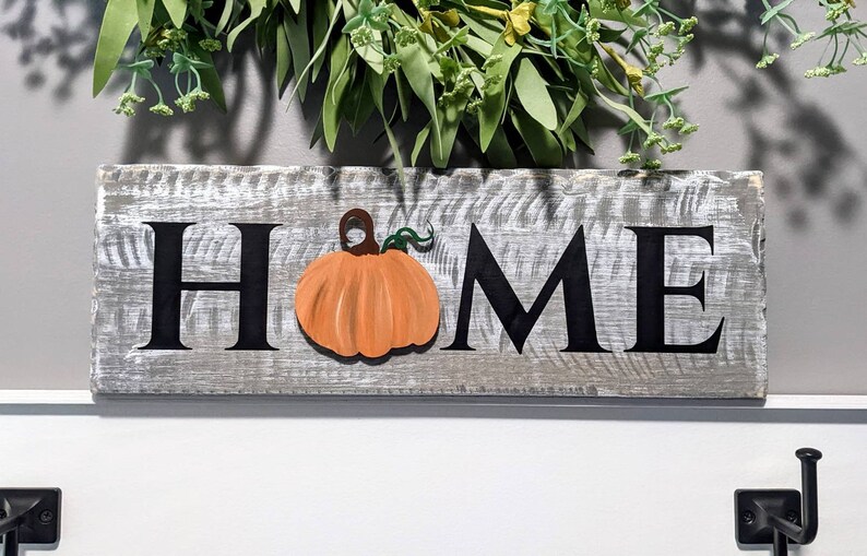 May include: A rustic wooden sign with the word "HOME" in black letters. An orange pumpkin is affixed to the sign. The sign is painted gray with white accents. Greenery is visible above the sign.