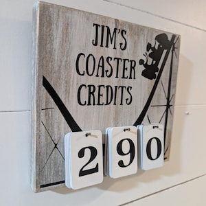 May include: A wooden sign with the text "JIM'S COASTER CREDITS" and a roller coaster graphic. Below the text are three white number tiles displaying "290". The sign has a distressed, gray and white finish.
