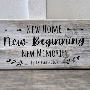 May include: White wooden sign with black text that reads "New Home New Beginning New Memories Established 2024". The sign has a distressed look and features arrows and leaves.