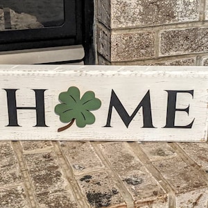May include: A rectangular wooden sign with the word "HOME" in black letters. A green shamrock is placed between the letters "M" and "E". The sign has a distressed, white-painted finish.
