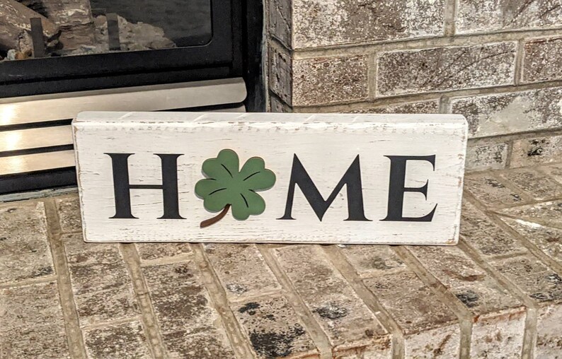 May include: A rectangular wooden sign with the word "HOME" in black letters. A green shamrock is placed between the letters "M" and "E". The sign has a distressed, white-painted finish.