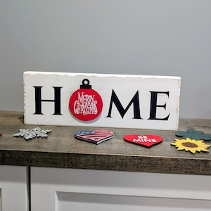May include: A white wooden sign with the word "HOME" in black letters. The "O" is replaced with a red Christmas ornament. Decorative wooden cutouts of a snowflake, heart, and sunflower are in front of the sign.