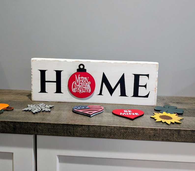 May include: A white wooden sign with the word "HOME" in black letters. The "O" is replaced with a red Christmas ornament. Decorative wooden cutouts of a snowflake, heart, and sunflower are in front of the sign.